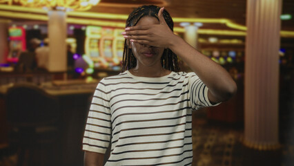 Woman covering eyes with hand and smiling at casino slot machines on indoor gaming floor; playful...
