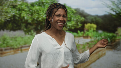 Fototapeta premium Woman with braided hair laughing, hand raised and dancing in a green forest garden outdoors near raised beds; happiness playful.