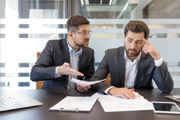 Two stressed businessmen discussing documents in an office, one man actively expressing disagreement and frustration while the other listens with a pensive and disappointed expression