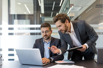 Two professional businessmen are collaborating and discussing work using a laptop and documents during a corporate meeting in a modern office environment, focusing on teamwork and partnership