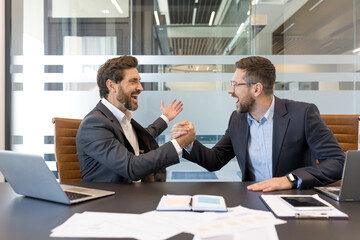 Two excited businessmen shaking hands in an arm wrestling grip, sitting at a conference table, celebrating success and achievement in their corporate office setting