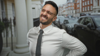 Naklejka premium Young man with beard grimacing in pain on urban street in a city setting, wearing glasses, smart attire, amidst parked cars and historical buildings in urban surroundings.