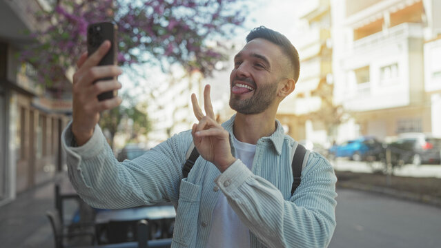 Smiling young man taking selfie with peace sign outdoors on sunny day, capturing joyful moment in lively urban street setting, showcasing youthful energy and modern lifestyle.