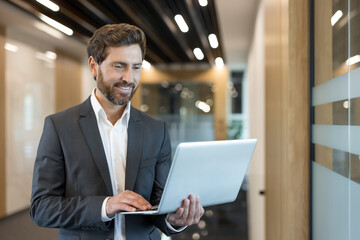 Fototapeta premium Confident businessman standing in a contemporary corporate corridor, smiling while using a laptop, representing work, technology, and success in a professional environment