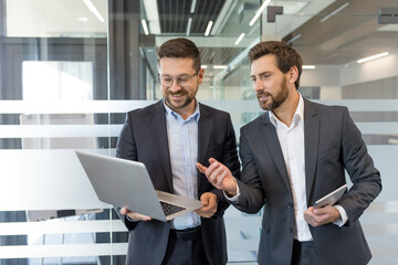 Two smiling businessmen in suits collaborate in a modern office, pointing at a laptop while one holds a tablet, discussing strategy, data and planning for corporate success
