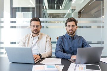 Two professional businessmen sitting at a modern office desk, actively working on their laptops and...
