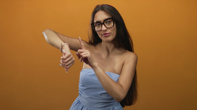 Woman in strapless blue dress and glasses pointing to watch on her wrist in an orange studio; impatience punctuality time awareness.