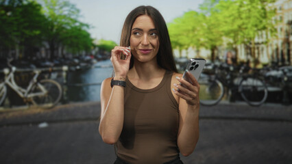 Fototapeta premium Woman holding smartphone and waving hand on amsterdam street beside canal; travel connection serenity.