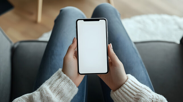 Smartphone with white screen mockup in the hands of a woman sitting on the sofa at home