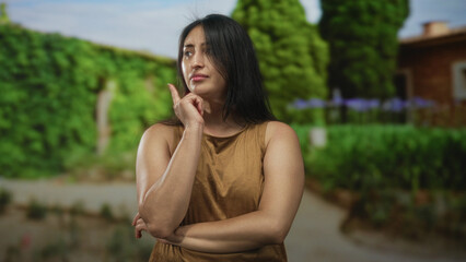 Woman with finger to nose and arms crossed in a forest pathway, green outdoors setting; thoughtful...