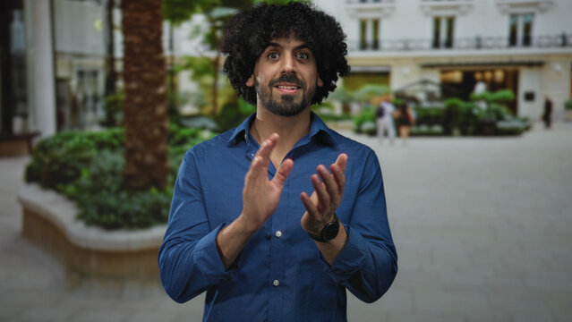 Man with curly hair clapping hands outdoors in front of a building and plaza, smiling while applauding; applause celebration.