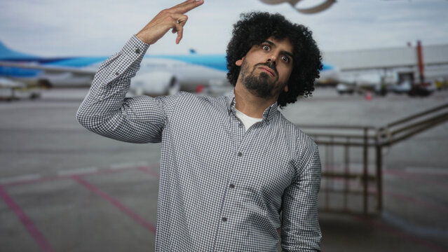 Man with curly hair points finger upward at airport terminal with raised hand and intense wide eyes, wearing checked shirt; travel fatigue. - Powered by Adobe