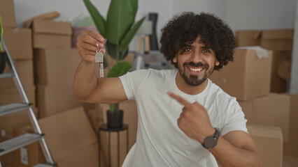 Man holding house keys and pointing finger to keys while smiling inside building surrounded by moving boxes; new home excitement.