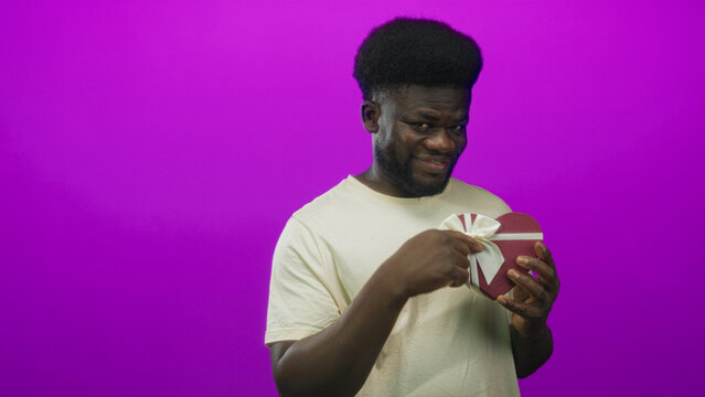Man points finger to heart box with ribbon while holding it against a magenta studio wall; playful flirtation charm. - Powered by Adobe