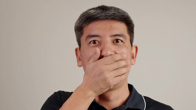 Surprised brunette man covering his mouth with his hand, showing shock, disbelief, or sudden realization on clean neutral studio background.