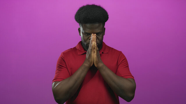 Man with hands pressed together in prayer, eyes closed and wearing a red polo, standing before a purple studio wall; contemplation.