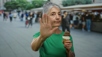 Senior woman with grey hair at an outdoor market holding an ice cream cone gestures stop,...