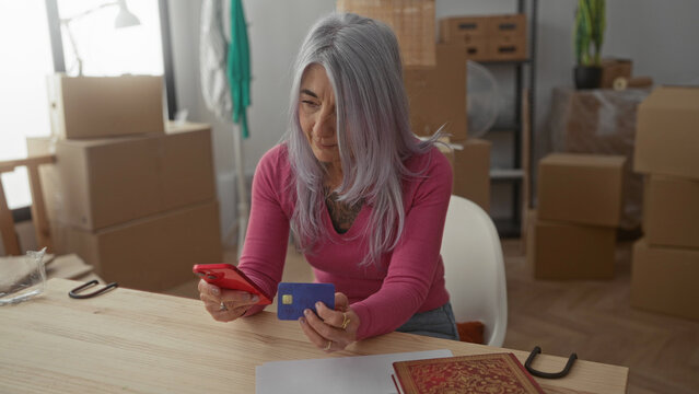Senior woman in new home living room holding smartphone and credit card with unpacked boxes in the background, portraying an indoor modern lifestyle.