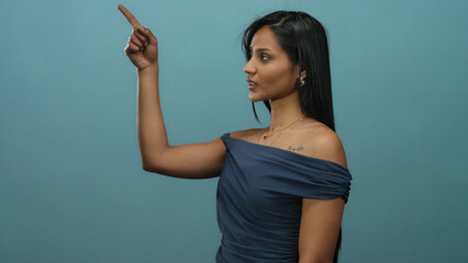 Young latin woman in a blue dress gesturing with one finger up against an isolated blue background...