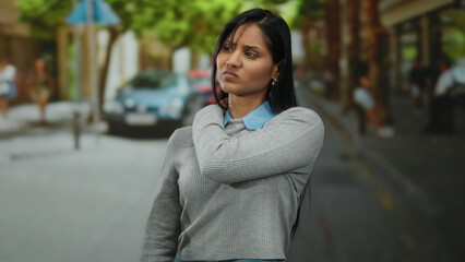 Fototapeta premium Young woman in gray sweater holds shoulder in discomfort on urban street with blurred background and trees lining the sidewalk.