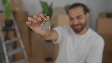 Man holds house keyring and keys in house with hand extended toward camera, moving boxes behind him; new home happiness.