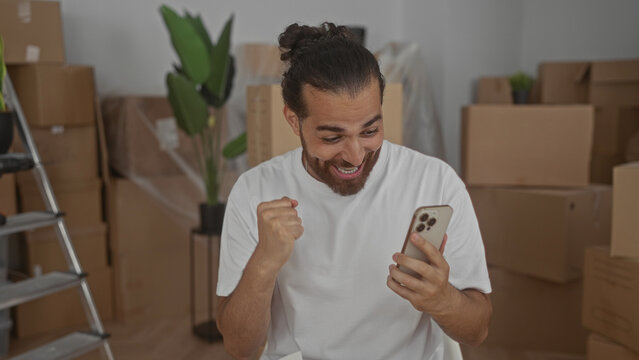 Man holding smartphone, fist pump gesture in building amid packed boxes and ladder; moving day excitement. - Powered by Adobe