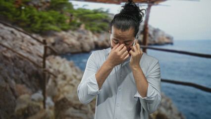 Man holding smartphone to ear and rubbing forehead on seaside street by rocky coast and wooden railing in rolled sleeves; private worry.