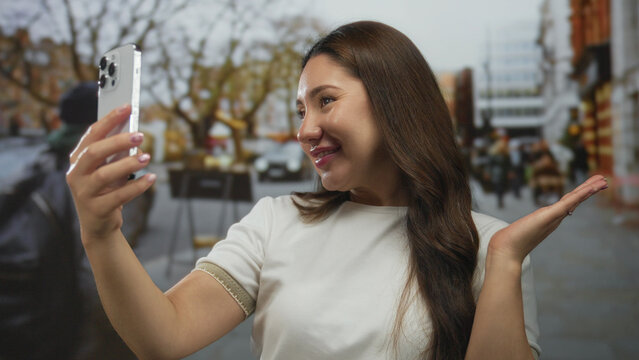 Hispanic woman holding smartphone and raising hand while taking a selfie on a busy city street; social connection joy. - Powered by Adobe