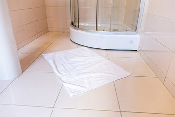 White foot towel on pale tile floor beside a curved glass shower enclosure. Bathroom, hygiene, everyday home comfort.