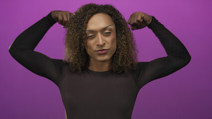 Hispanic transgender woman in black top flexes arms with clenched fists and confident smile in purple studio; pride.