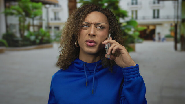 Woman holds smartphone to ear with raised hand and furrowed brow on urban city street in front of building; frustration concern tension. - Powered by Adobe