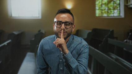 Man wearing glasses and denim shirt finger to lips for silence in church building; reflection faith...