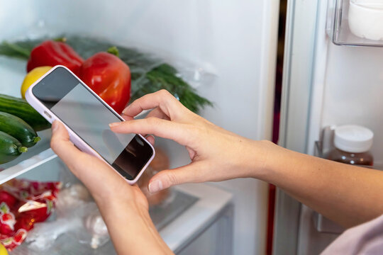 Woman using smartphone to take photo of fresh vegetables in refrigerator, showcasing healthy eating habits and modern technology in kitchen environment with vibrant colors