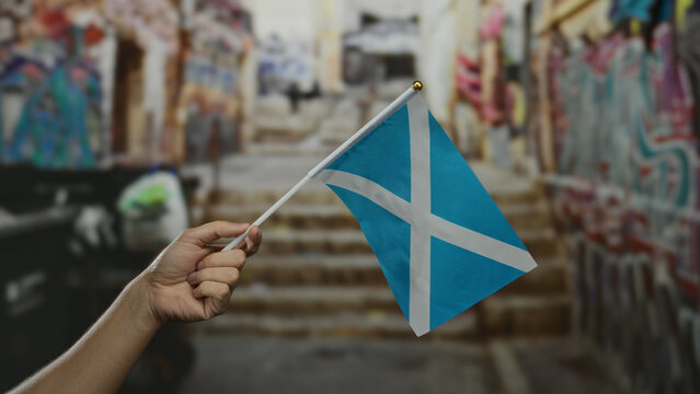 Hand holding scottish flag in urban street scene with colorful graffiti on stairs, evoking cultural pride and vibrant city atmosphere.