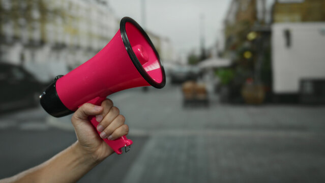 Man holding pink megaphone in city street signifies public announcement, showcasing hand raised with blurred background indicating urban action and spontaneous speech.