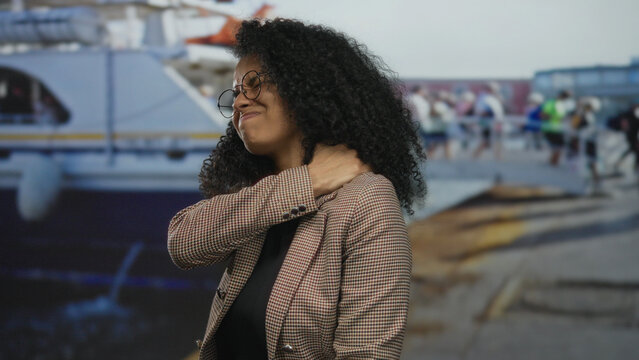 Young hispanic woman with curly hair and glasses holds neck in pain at port dock on a boat quay; discomfort.