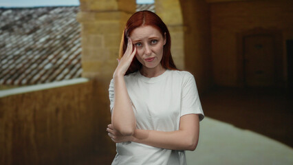 Woman standing in outdoor university courtyard with hand on head, appearing thoughtful and reflective, showcasing a serene and picturesque academic environment.
