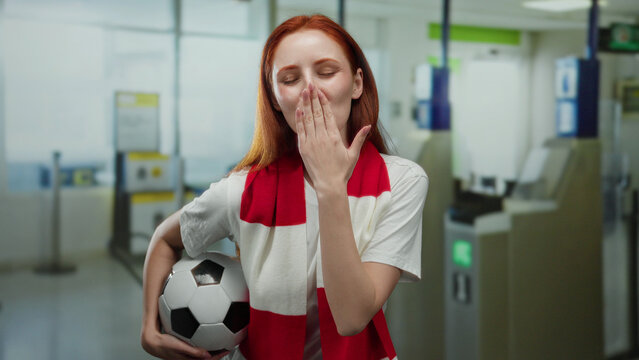 Woman with red hair holding soccer ball at airport terminal, smiling and making a hand gesture, wearing a red and white scarf, capturing a moment of excitement and anticipation. - Powered by Adobe