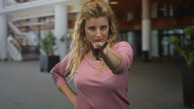 Woman points finger at camera with hand raised in building atrium, arm bent and palm visible near face; confidence defiance.