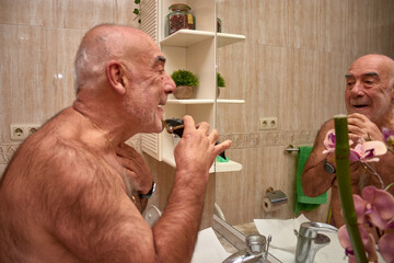 A focused senior citizen cuts his hair and trims his beard in front of the mirror in a simply...