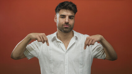 Young hispanic man performs double finger pointing gesture downward in red studio; self confidence growth.