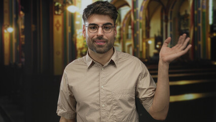 Man in beige shirt and round glasses smiling and waving hand in church building; serenity faith.