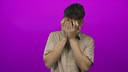 Young hispanic man with glasses and beige shirt holding hands under chin in purple studio; doubt...