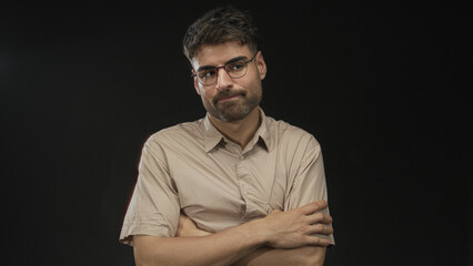 Young hispanic man with glasses crosses arms in studio under black lighting wearing beige shirt; confidence.