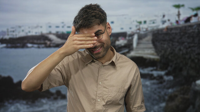 Hispanic man in beige shirt covers his eyes with hand beside a rocky seaside building on stone pier walkway; solitude reflection. - Powered by Adobe
