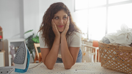 Woman with hands on cheeks leaning on ironing board with iron and laundry basket in house; home...