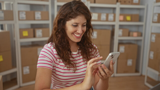 Smiling woman taps smartphone with finger and makes a fist pump in a storage building among cardboard parcels on shelves; joy success.