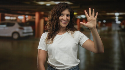 Woman wearing white tshirt holds up open hand beside parked car in parking building smiling at camera  friendly greeting. © Krakenimages.com