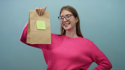 Woman holding a brown paper bag with green sticky note and smiley face in blue studio while smiling...