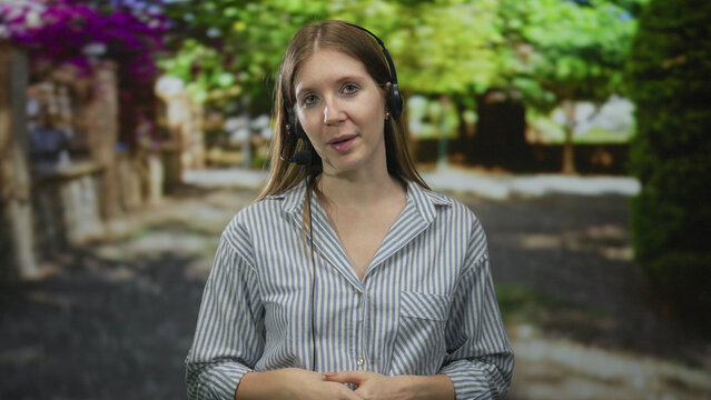 Woman speaking into microphone headset with hand gesture amid lush tree foliage and sunlit green pathway in forest; confidence.